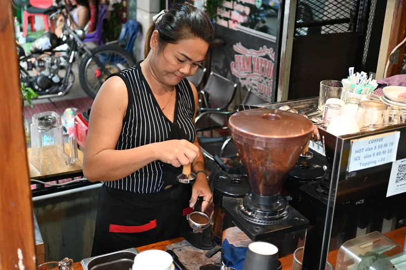 A barista holds a portafilter and tamper beside a coffee grinder at a street-side counter